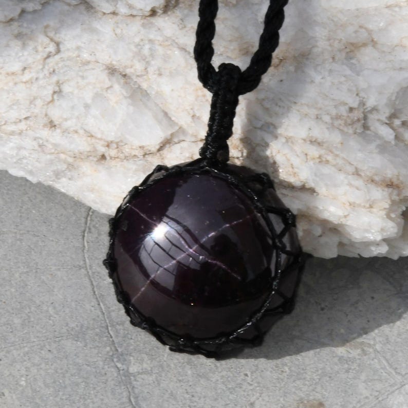 A single, smooth, polished, oval Star Garnet necklace pendant is displayed on a textured light rock surface. The dark red to reddish-purple gemstone exhibits a distinct four-rayed star (asterism) in its center when light reflects off it. It is encased in a black macrame net that forms the necklace cord. Star Garnet is associated with inner strength, protection, vitality, and regeneration.