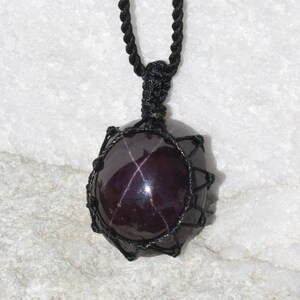 Close-up of a polished star garnet pendant in deep burgundy, wine-red, and dark plum tones, displayed on a light stone background. A distinct four-rayed star appears across the gemstone when illuminated, creating a bright reflective cross. The garnet has a smooth glossy finish with rich color depth and natural translucency. It is secured in a black macrame net and attached to a black adjustable cord. Associated with passion, vitality, grounding, courage, and emotional strength.