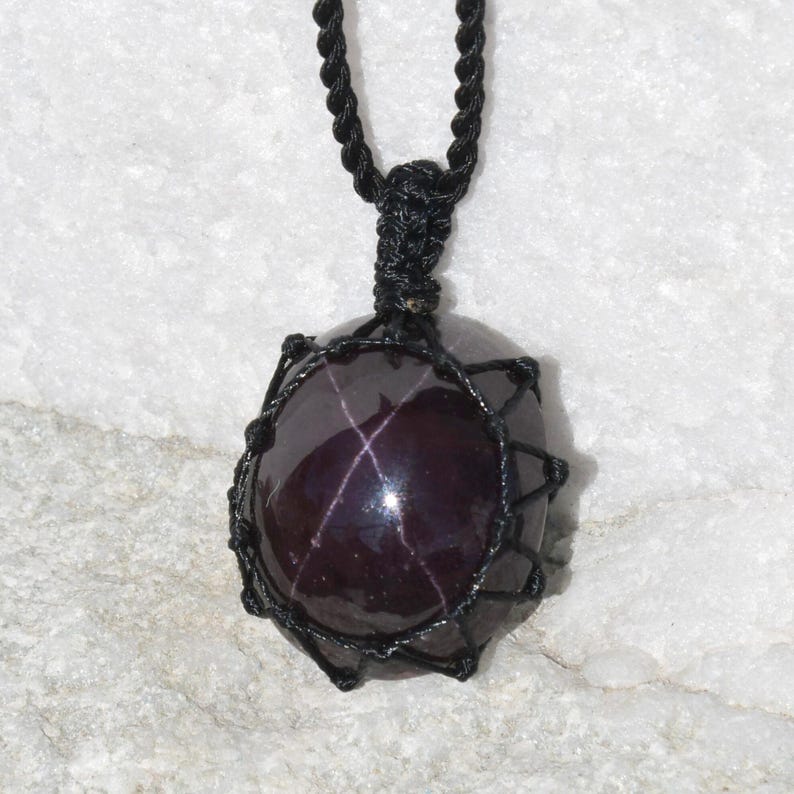 Close-up of a polished star garnet pendant in deep burgundy, wine-red, and dark plum tones, displayed on a light stone background. A distinct four-rayed star appears across the gemstone when illuminated, creating a bright reflective cross. The garnet has a smooth glossy finish with rich color depth and natural translucency. It is secured in a black macrame net and attached to a black adjustable cord. Associated with passion, vitality, grounding, courage, and emotional strength.
