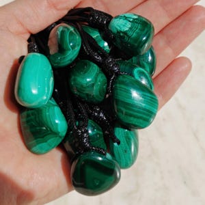 Close-up of multiple polished irregular Malachite in shades of vivid green and deep forest green, held in a hand against a light background. The green opaque gemstones have smooth glossy surfaces with distinct banded patterns, concentric rings, and flowing lines, with varied shapes and layered green formations visible across the stones. It is attached to a black cord with a top knot and adjustable knot closure. Malachite is associated with transformation, protection, and emotional healing.