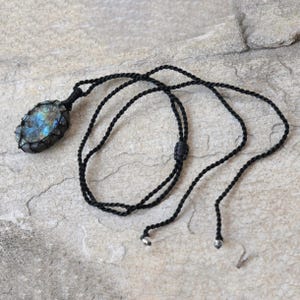 Raw Labradorite pendant displaying vivid flashes of electric blue, turquoise, gold, and hints of green across its natural rough surface. The stone is wrapped in a black macrame net with an adjustable black cord with a sliding knot closure and crystal beads and displayed on a light stone background. Labradorite is associated with transformation, intuition, protection, inner strength, spiritual awakening,  and enhanced creativity.
