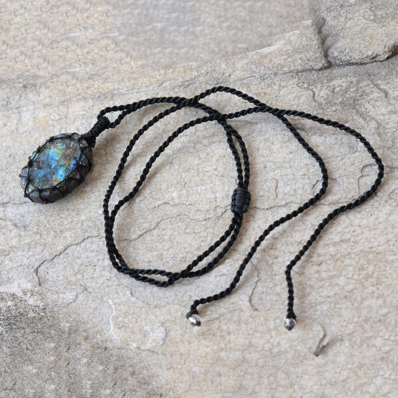 Raw Labradorite pendant displaying vivid flashes of electric blue, turquoise, gold, and hints of green across its natural rough surface. The stone is wrapped in a black macrame net with an adjustable black cord with a sliding knot closure and crystal beads and displayed on a light stone background. Labradorite is associated with transformation, intuition, protection, inner strength, spiritual awakening,  and enhanced creativity.