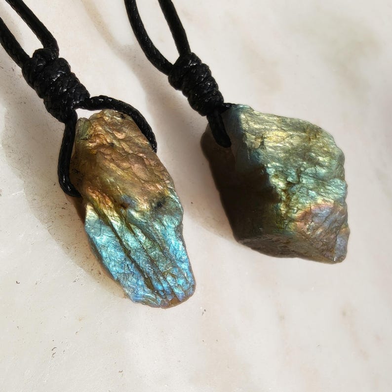 Close-up of two raw irregular Labradorite in shades of blue, teal, green and golden brown, resting on a light stone surface. The Labradorite gemstones have a natural rough texture with strong iridescent flashes, visible striations, and layered color play with reflective highlights. They are secured with a top knot and attached to black cords with adjustable knot closure. Labradorite is associated with transformation, protection, intuition, and inner strength.