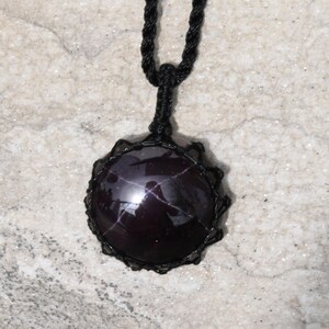 A single, smooth, polished, oval Star Garnet necklace pendant is displayed on a textured light rock surface. The dark red to reddish-purple gemstone exhibits a distinct four-rayed star (asterism) in its center when light reflects off it. It is encased in a black macrame net that forms the necklace cord. Star Garnet is associated with inner strength, protection, vitality, and regeneration.