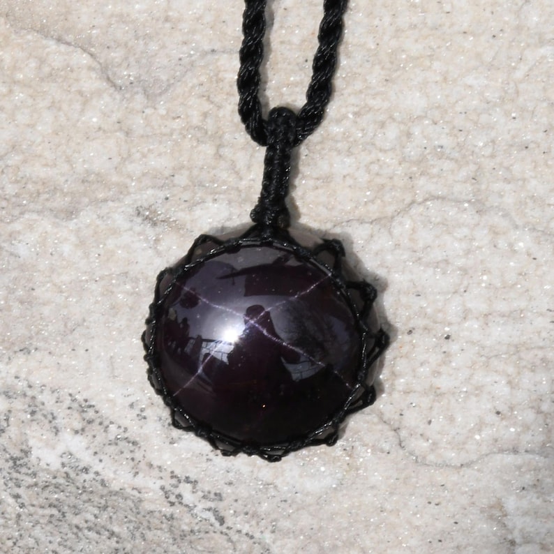 A single, smooth, polished, oval Star Garnet necklace pendant is displayed on a textured light rock surface. The dark red to reddish-purple gemstone exhibits a distinct four-rayed star (asterism) in its center when light reflects off it. It is encased in a black macrame net that forms the necklace cord. Star Garnet is associated with inner strength, protection, vitality, and regeneration.