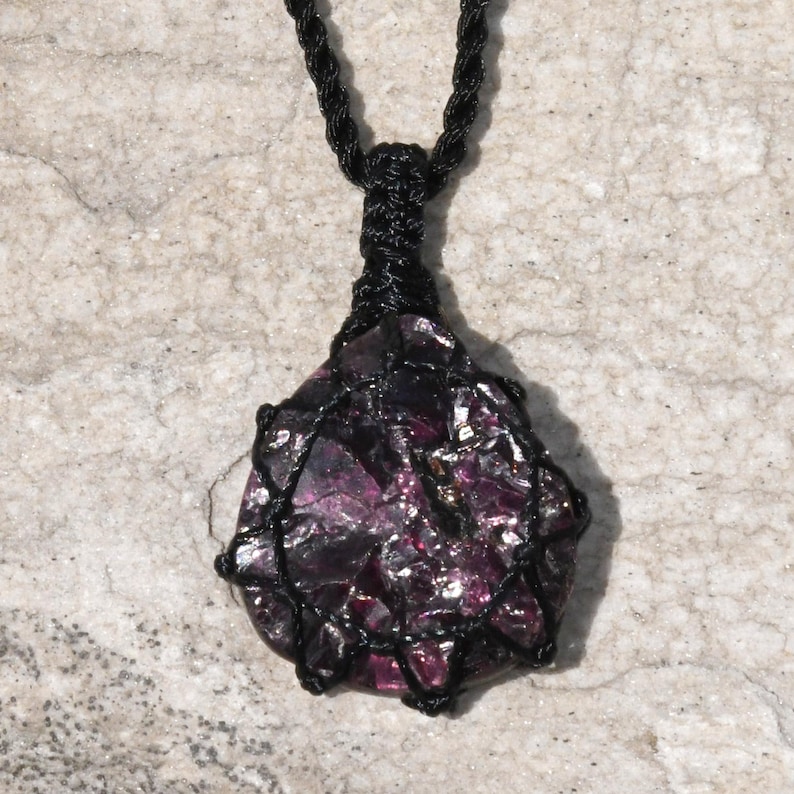 A single, raw, dtop shaped raw Garnet necklace pendant is displayed on a textured white rock surface. The dark reddish-purple gemstone has a rough, unpolished texture. It is encased in a black macrame net that forms the necklace cord. Garnet is associated with passion, energy, protection, and healing.