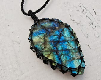 Puede incluir: Un colgante de labradorita en forma de lágrima con destellos iridiscentes azules y verdes, encerrado en una red de cordón de macramé negro. La piedra cuelga de un collar de cordón trenzado negro a juego, mostrando una superficie natural texturizada.