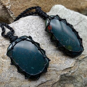 Two Bloodstone pendant necklaces displayed against a textured natural stone attached to adjustable twisted black cords with a sliding knot closure. Each polished oval gemstone features deep forest-green tones with subtle natural red mineral inclusions, making every pendant unique. Bloodstone is associated with Courage, Resilience, Grounding, Vitality, and Inner Strength.