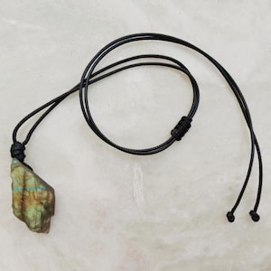 Close-up of a raw irregular Labradorite in shades of green and golden brown, resting on a light stone surface with the full black cord visible. The Labradorite gemstone has a natural rough texture with strong iridescent flashes, visible striations, and layered color play with reflective highlights. It is secured with a top knot and attached to a black cord with adjustable knot closure. Labradorite is associated with transformation, protection, intuition, and inner strength.