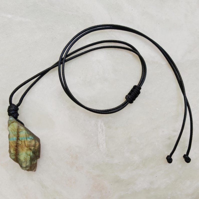 Close-up of a raw irregular Labradorite in shades of green and golden brown, resting on a light stone surface with the full black cord visible. The Labradorite gemstone has a natural rough texture with strong iridescent flashes, visible striations, and layered color play with reflective highlights. It is secured with a top knot and attached to a black cord with adjustable knot closure. Labradorite is associated with transformation, protection, intuition, and inner strength.