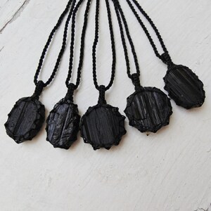 Close-up of 5 raw oval and round Black Tourmaline in deep black shades, resting on a white wood surface. The Black Tourmaline dark gemstone has strong parallel vertical striations, rough uneven edges, and a matte to slightly reflective texture with natural ridges and occasional lighter inclusions and surface variations. Each piece is wrapped in a black macrame net and attached to a black cord with an adjustable knot closure. Black Tourmaline is associated with grounding and protection.