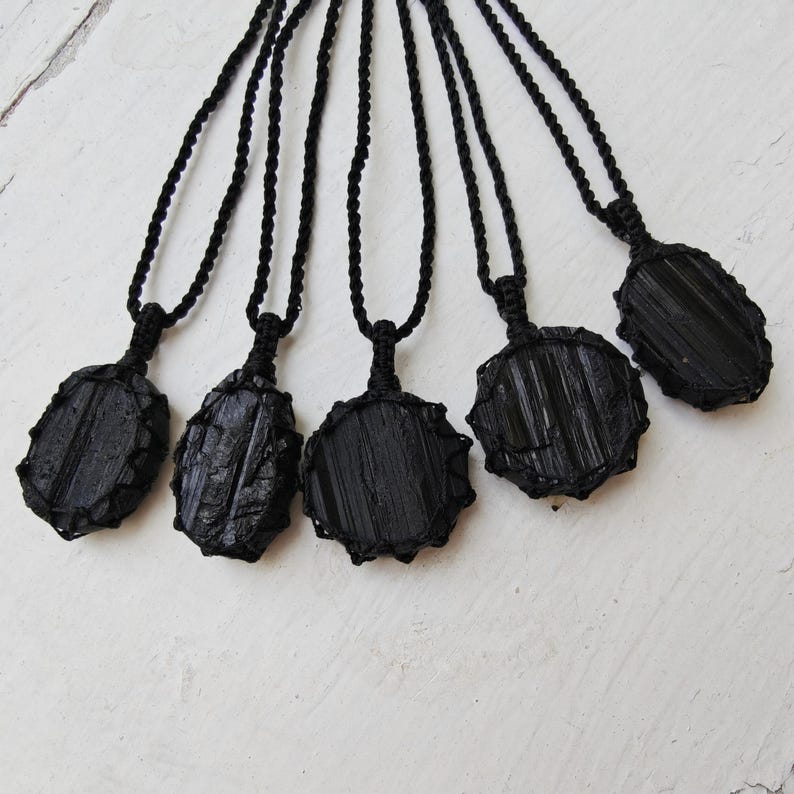 Close-up of 5 raw oval and round Black Tourmaline in deep black shades, resting on a white wood surface. The Black Tourmaline dark gemstone has strong parallel vertical striations, rough uneven edges, and a matte to slightly reflective texture with natural ridges and occasional lighter inclusions and surface variations. Each piece is wrapped in a black macrame net and attached to a black cord with an adjustable knot closure. Black Tourmaline is associated with grounding and protection.