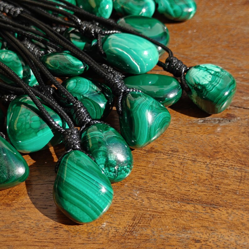 Close-up of multiple polished irregular Malachite in shades of vivid green and deep forest green, clustered together against a textured wood background. The green opaque gemstones have smooth glossy surfaces with distinct banded patterns, flowing concentric lines, and eye-like formations with darker green swirls. It is attached to a black cord with a top knot and adjustable knot closure. Malachite is associated with transformation, protection, and emotional healing.