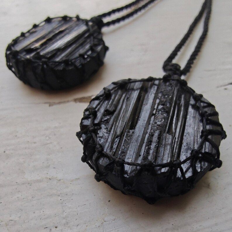 Close-up of 2 raw round Black Tourmaline in deep black shades, resting on a white wood surface. The Black Tourmaline dark gemstone has strong parallel vertical striations, rough uneven edges, and a matte to slightly reflective texture with natural ridges and occasional lighter inclusions and surface variations. Each piece is wrapped in a black macrame net and attached to a black cord with an adjustable knot closure. Black Tourmaline is associated with grounding and protection.