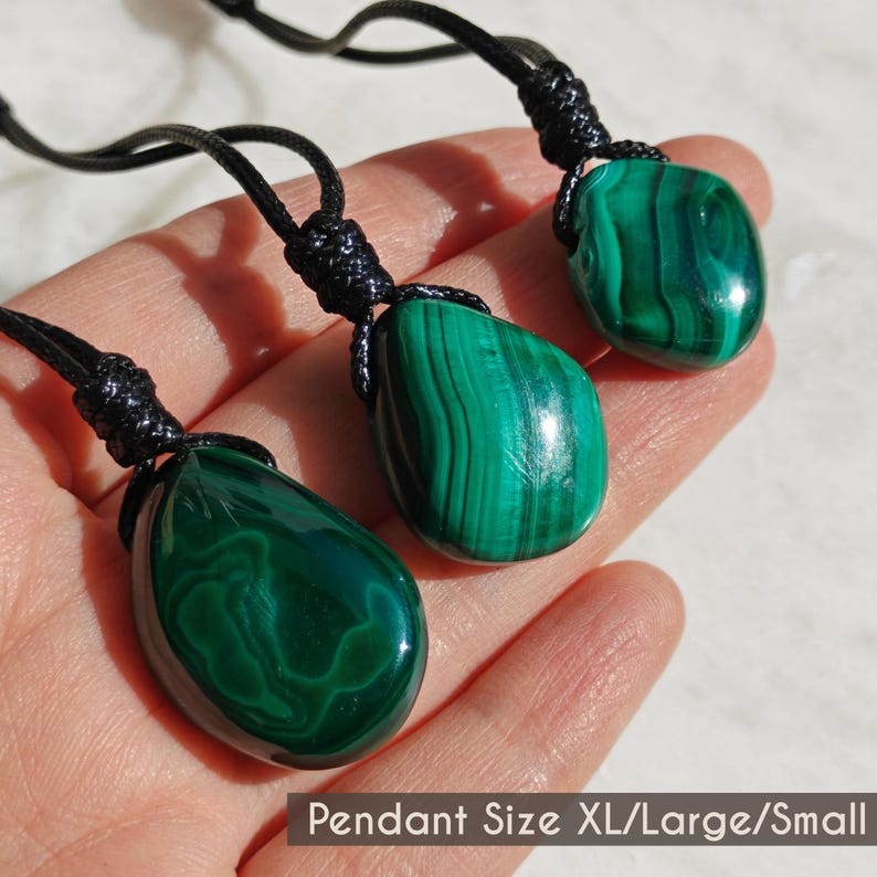 Close-up of 3 polished irregular Malachite in shades of vivid green and deep forest green, displayed on a hand against a light background. The green opaque gemstones have smooth glossy surfaces with distinct banded patterns, flowing lines, and concentric formations, with light reflections highlighting their curved shapes and surface polish. It is attached to a black cord with a top knot and adjustable knot closure. The image includes the text Pendant Size XL Large Small.
