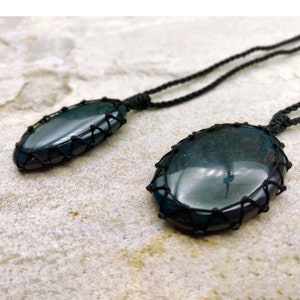 Two Bloodstone pendant necklaces displayed against a textured natural stone attached to adjustable twisted black cords with a sliding knot closure. Each polished oval gemstone features deep forest-green tones with subtle natural red mineral inclusions, making every pendant unique. Bloodstone is associated with Courage, Resilience, Grounding, Vitality, and Inner Strength.