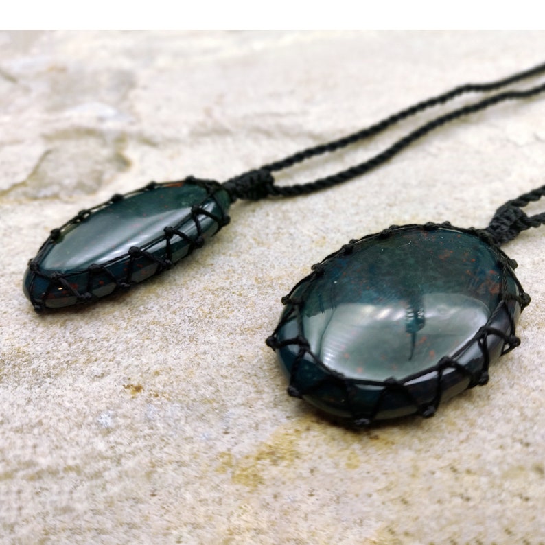 Two Bloodstone pendant necklaces displayed against a textured natural stone attached to adjustable twisted black cords with a sliding knot closure. Each polished oval gemstone features deep forest-green tones with subtle natural red mineral inclusions, making every pendant unique. Bloodstone is associated with Courage, Resilience, Grounding, Vitality, and Inner Strength.