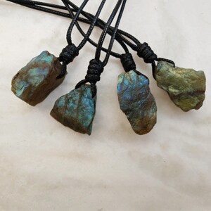 Close-up of 4 raw irregular Labradorite in shades of blue, teal, and golden brown, resting on a light stone surface. The Labradorite gemstones have a natural rough texture with strong iridescent flashes, visible striations, and layered color play with reflective highlights. They are secured with a top knot and attached to black cords with adjustable knot closure. Labradorite is associated with transformation, protection, intuition, and inner strength.