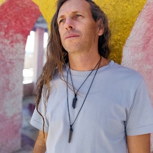 a man wearing two-layered necklaces with dark, pointed pendants against a brightly colored wall. The pendants are elongated, transparent brown/smoky Smoky Quartz crystals. Smoky Quartz is associated with grounding, protection, and emotional calmness.
