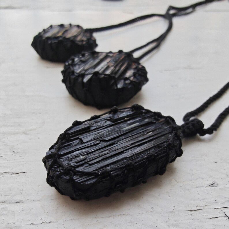 Close-up of 3 raw oval and round Black Tourmaline in deep black shades, resting on a white wood surface. The Black Tourmaline dark gemstone has strong parallel vertical striations, rough uneven edges, and a matte to slightly reflective texture with natural ridges and occasional lighter inclusions and surface variations. Each piece is wrapped in a black macrame net and attached to a black cord with an adjustable knot closure. Black Tourmaline is associated with grounding and protection.