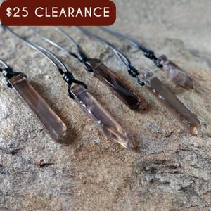 Five raw smoky quartz necklaces with rectangular stones in dark brown translucent hues, rough-textured with irregular edges, each hung on a knotted black cord, placed on a light brown stone surface with a natural grainy texture. Smokey Quartz is associated with grounding, protection, and emotional calmness.