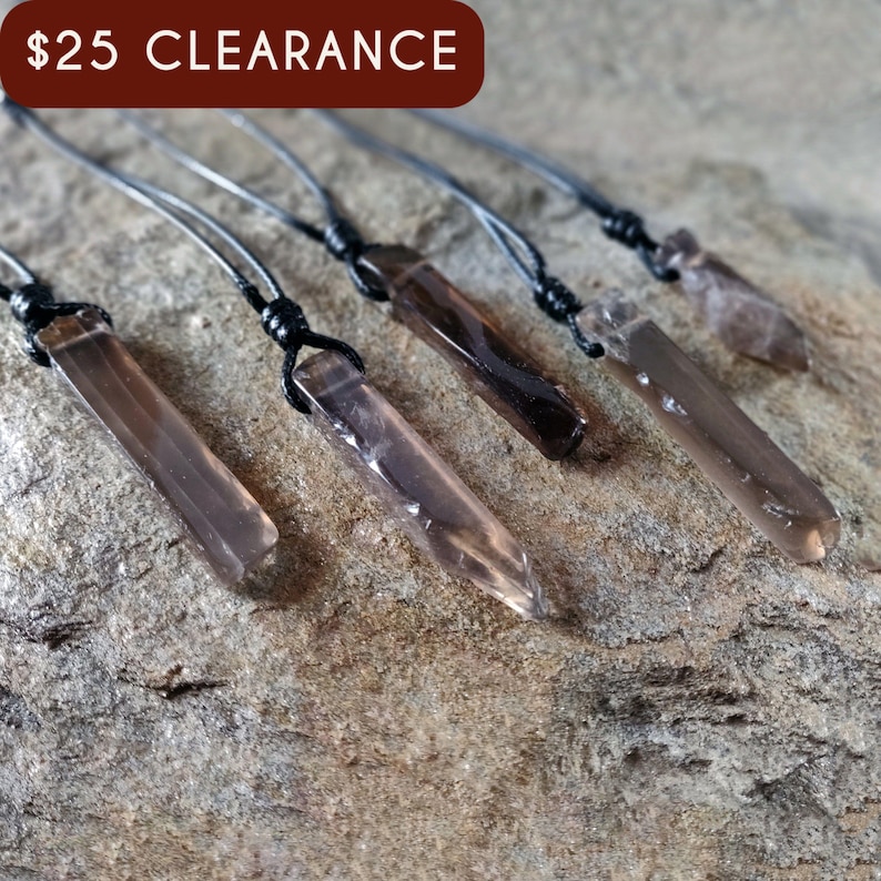 Five raw smoky quartz necklaces with rectangular stones in dark brown translucent hues, rough-textured with irregular edges, each hung on a knotted black cord, placed on a light brown stone surface with a natural grainy texture. Smokey Quartz is associated with grounding, protection, and emotional calmness.