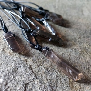 Few raw smoky quartz necklaces with rectangular stones in dark brown translucent hues, rough-textured with irregular edges, each hung on a knotted black cord, placed on a light brown stone surface with a natural grainy texture. Smokey Quartz is associated with grounding, protection, and emotional calmness.
