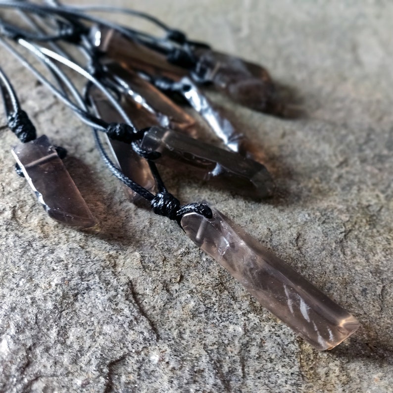 Few raw smoky quartz necklaces with rectangular stones in dark brown translucent hues, rough-textured with irregular edges, each hung on a knotted black cord, placed on a light brown stone surface with a natural grainy texture. Smokey Quartz is associated with grounding, protection, and emotional calmness.