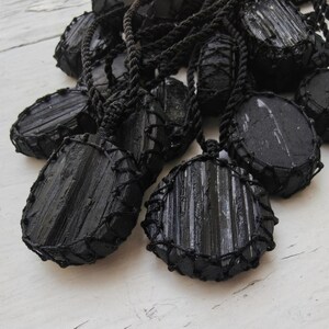 Close-up of multiple raw oval and round Black Tourmaline in deep black shades, resting on a white wood surface. The Black Tourmaline dark gemstone has strong parallel vertical striations, rough uneven edges, and a matte to slightly reflective texture with natural ridges and occasional lighter inclusions and surface variations. Each piece is wrapped in a black macrame net and attached to a black cord with an adjustable knot closure. Black Tourmaline is associated with grounding and protection.