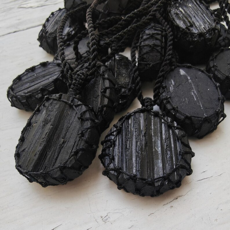 Close-up of multiple raw oval and round Black Tourmaline in deep black shades, resting on a white wood surface. The Black Tourmaline dark gemstone has strong parallel vertical striations, rough uneven edges, and a matte to slightly reflective texture with natural ridges and occasional lighter inclusions and surface variations. Each piece is wrapped in a black macrame net and attached to a black cord with an adjustable knot closure. Black Tourmaline is associated with grounding and protection.