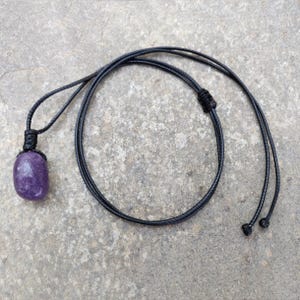May include: A purple gemstone pendant necklace with a black cord. The gemstone is a rounded rectangle shape. The cord is knotted at the top of the pendant and has adjustable sliding knots at the ends.