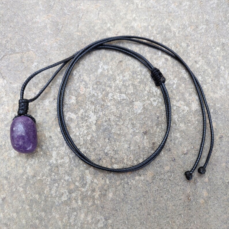 May include: A purple gemstone pendant necklace with a black cord. The gemstone is a rounded rectangle shape. The cord is knotted at the top of the pendant and has adjustable sliding knots at the ends.