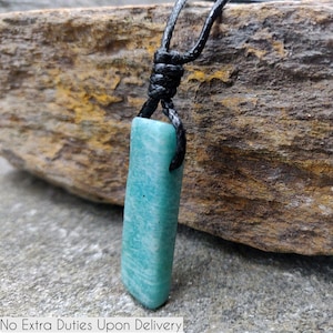 May include: A rectangular, light blue pendant necklace with a black cord. The pendant is a smooth, polished stone with a marbled appearance. The necklace is displayed against a textured, neutral-toned background. The text "No Extra Duties Upon Delivery" is at the bottom.