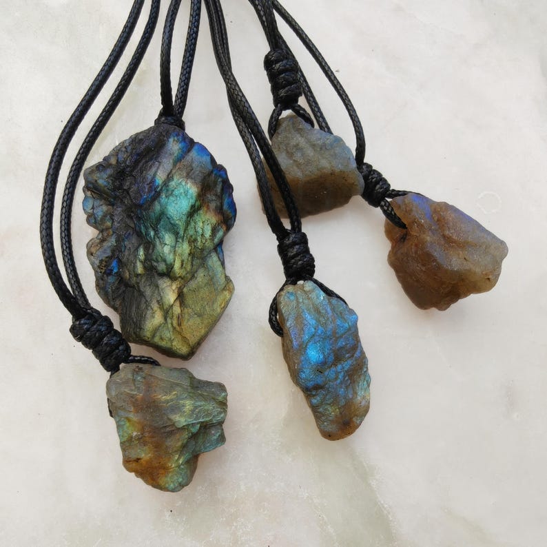 Close-up of 5 raw irregular Labradorite stones in shades of blue, teal, green, and golden hues, displayed on a light surface. The Labradorite gemstones have a rough natural texture with strong iridescent flashes, layered color play, and visible striations across their surfaces. Each stone is secured with a top knot and attached to a black cord with adjustable knot closure. Labradorite is associated with transformation, protection, intuition, and spiritual shielding.