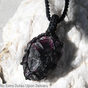 May include: A dark, rough-textured gemstone pendant with a deep burgundy interior, encased in a black woven macrame cord. The pendant hangs from a matching black cord. The text "No Extra Duties Upon Delivery" is visible at the bottom.