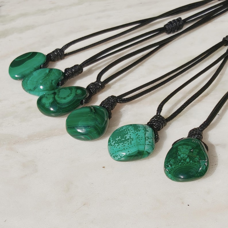 Close-up of 6 polished irregular Malachite in shades of vivid green and deep forest green, arranged on a light stone surface. The green opaque gemstones have smooth glossy surfaces with distinct banded patterns, concentric lines, and varied formations, including rounded and triangular shapes with layered green stripes and darker swirls. It is attached to a black cord with a top knot and adjustable knot closure. Malachite is associated with transformation, protection, and emotional healing.