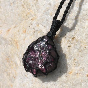 A single, raw, dtop shaped raw Garnet necklace pendant is displayed on a textured white rock surface. The dark reddish-purple gemstone has a rough, unpolished texture. It is encased in a black macrame net that forms the necklace cord. Garnet is associated with passion, energy, protection, and healing.