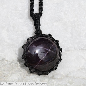 A single, smooth, polished, oval Star Garnet necklace pendant is displayed on a textured light rock surface. The dark red to reddish-purple gemstone exhibits a distinct four-rayed star (asterism) in its center when light reflects off it. It is encased in a black macrame net that forms the necklace cord. Star Garnet is associated with inner strength, protection, vitality, and regeneration.