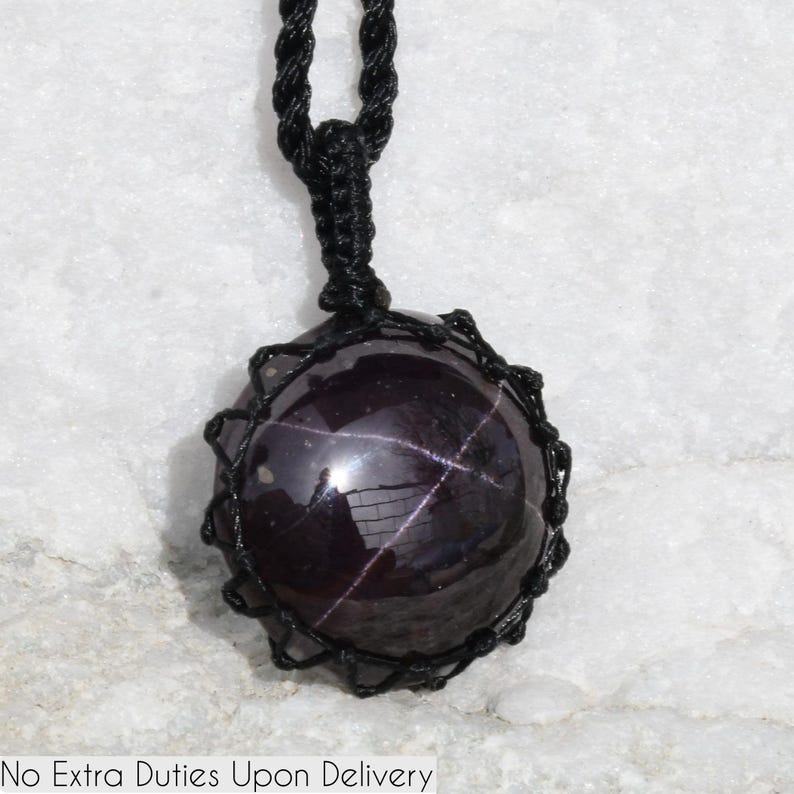 A single, smooth, polished, oval Star Garnet necklace pendant is displayed on a textured light rock surface. The dark red to reddish-purple gemstone exhibits a distinct four-rayed star (asterism) in its center when light reflects off it. It is encased in a black macrame net that forms the necklace cord. Star Garnet is associated with inner strength, protection, vitality, and regeneration.