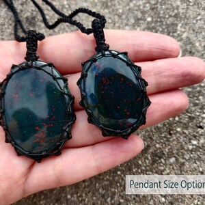 Two Bloodstone pendant necklaces displayed on a hand against a textured natural stone attached to adjustable twisted black cords with a sliding knot closure. Each polished oval gemstone features deep forest-green tones with subtle natural red mineral inclusions, making every pendant unique. Bloodstone is associated with Courage, Resilience, Grounding, Vitality, and Inner Strength.