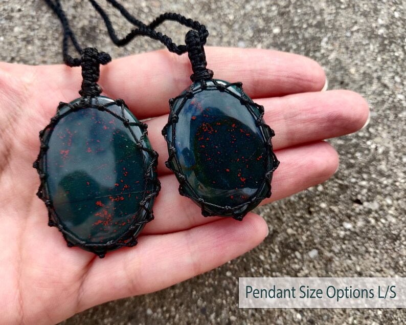 Two Bloodstone pendant necklaces displayed on a hand against a textured natural stone attached to adjustable twisted black cords with a sliding knot closure. Each polished oval gemstone features deep forest-green tones with subtle natural red mineral inclusions, making every pendant unique. Bloodstone is associated with Courage, Resilience, Grounding, Vitality, and Inner Strength.