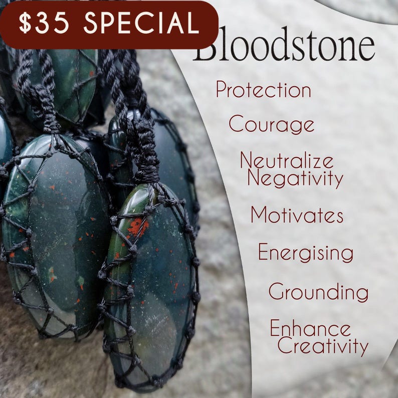Several Bloodstone pendant necklaces displayed together in black macrame settings, suspended from adjustable braided black cords against a natural stone background. The polished oval gemstones feature deep forest-green tones with scattered natural red mineral inclusions, making each pendant unique. The image includes the text "Bloodstone" and the qualities "Protection," "Courage," "Neutralize Negativity," "Motivates," "Energising," "Grounding," and "Enhance Creativity."