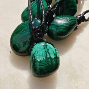 Close-up of multiple polished irregular Malachite in shades of vivid green and deep forest green, arranged on a light stone surface. The green opaque gemstones have smooth glossy surfaces with distinct banded patterns, concentric lines, and varied formations, including rounded and triangular shapes with layered green stripes and darker swirls. It is attached to a black cord with a top knot and adjustable knot closure. Malachite is associated with transformation, protection, and emotional healing