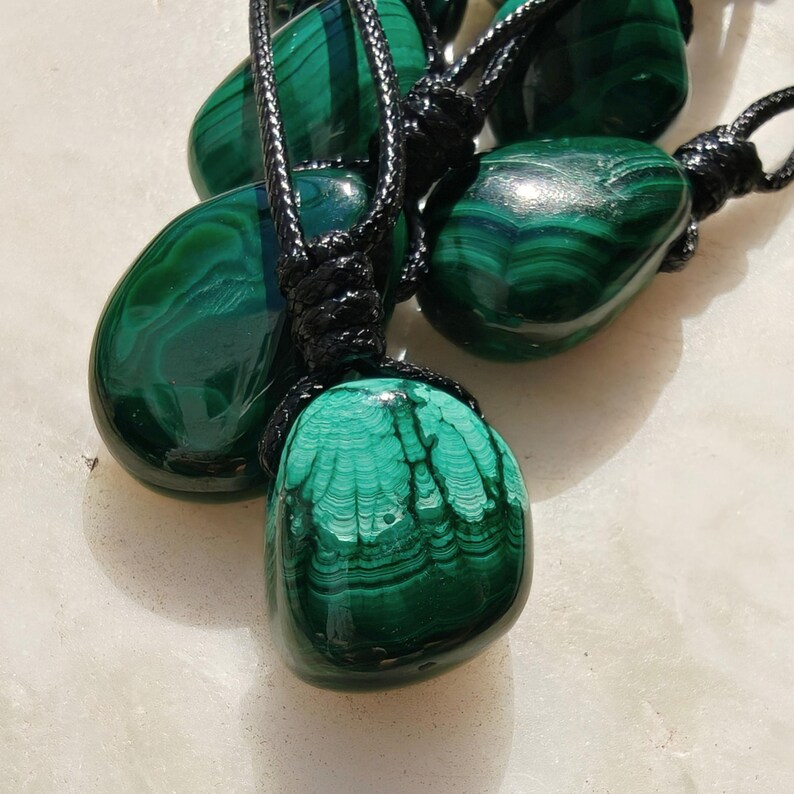 Close-up of multiple polished irregular Malachite in shades of vivid green and deep forest green, arranged on a light stone surface. The green opaque gemstones have smooth glossy surfaces with distinct banded patterns, concentric lines, and varied formations, including rounded and triangular shapes with layered green stripes and darker swirls. It is attached to a black cord with a top knot and adjustable knot closure. Malachite is associated with transformation, protection, and emotional healing