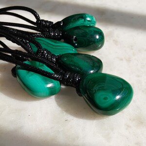 Close-up of multiple polished irregular Malachite in shades of vivid green and deep forest green, arranged on a light stone surface. The green opaque gemstones have smooth glossy surfaces with distinct banded patterns, concentric lines, and varied formations, including rounded and triangular shapes with layered green stripes and darker swirls. It is attached to a black cord with a top knot and adjustable knot closure. Malachite is associated with transformation, protection, and emotional healing