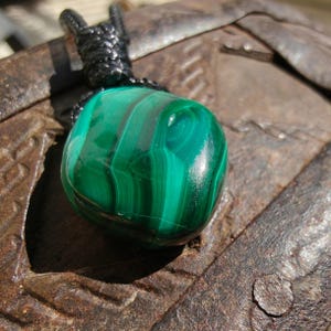 Close-up of a polished teardrop Malachite in shades of vivid green and deep forest green, resting on a textured brown surface. The green opaque gemstone has a smooth glossy surface with distinct banded patterns, flowing concentric lines, and layered swirls, with light reflections highlighting its curved form. It is attached to a black cord with a top knot and adjustable knot closure. Malachite is associated with transformation, protection, and emotional healing.