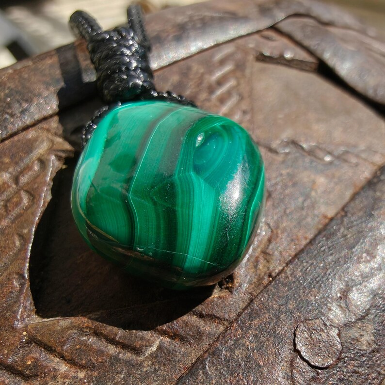 Close-up of a polished teardrop Malachite in shades of vivid green and deep forest green, resting on a textured brown surface. The green opaque gemstone has a smooth glossy surface with distinct banded patterns, flowing concentric lines, and layered swirls, with light reflections highlighting its curved form. It is attached to a black cord with a top knot and adjustable knot closure. Malachite is associated with transformation, protection, and emotional healing.