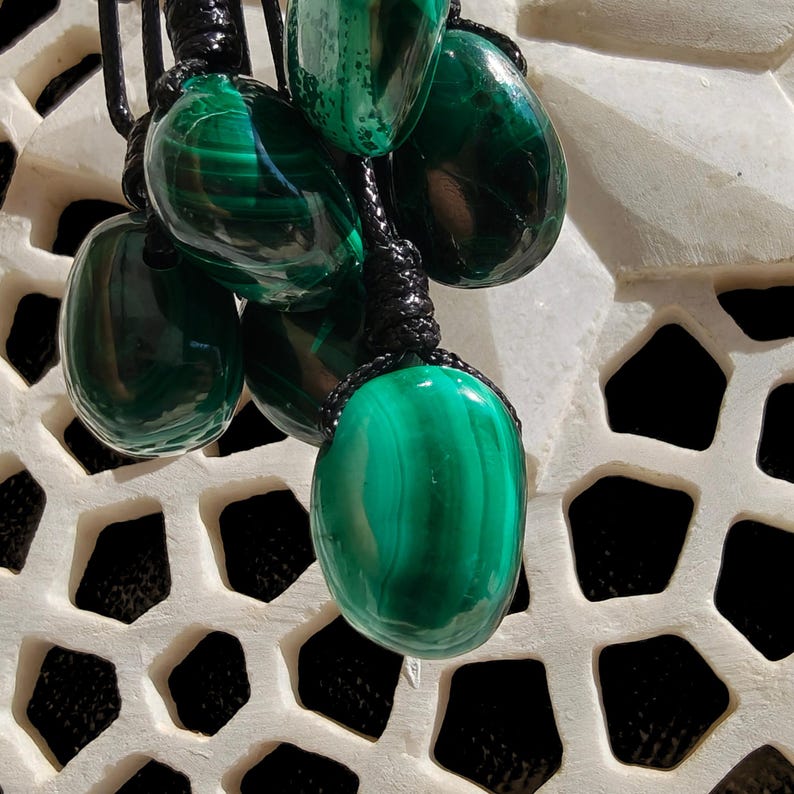 Multiple polished oval Malachite in shades of vivid green and deep forest green, hanging against a carved light stone surface with geometric openings. The green opaque gemstones have smooth glossy surfaces with distinct banded patterns, flowing lines, and layered green gradients, with light reflections and darker areas visible in overlapping pieces. It is attached to a black cord with a top knot. Malachite is associated with transformation, protection, and emotional healing.