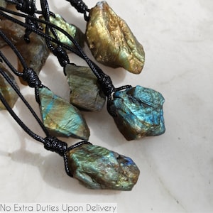Close-up of multiple raw irregular Labradorite stones in shades of blue, teal, green, and golden hues, displayed on a light surface. The Labradorite gemstones have a rough natural texture with strong iridescent flashes, layered color play, and visible striations across their surfaces. Each stone is secured with a top knot and attached to a black cord with adjustable knot closure. Labradorite is associated with transformation, protection, intuition, and spiritual shielding.
