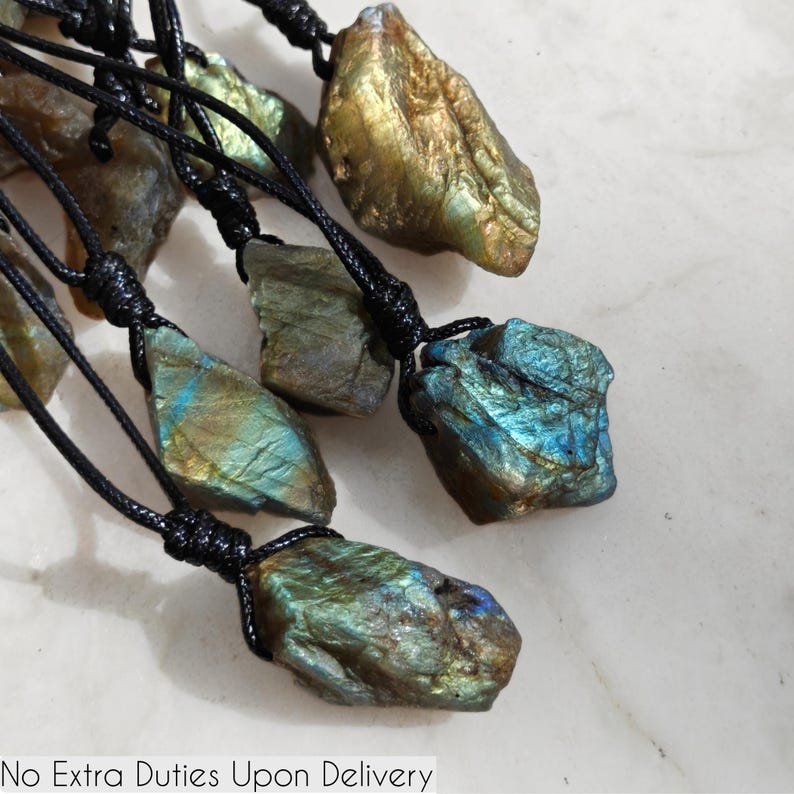 Close-up of multiple raw irregular Labradorite stones in shades of blue, teal, green, and golden hues, displayed on a light surface. The Labradorite gemstones have a rough natural texture with strong iridescent flashes, layered color play, and visible striations across their surfaces. Each stone is secured with a top knot and attached to a black cord with adjustable knot closure. Labradorite is associated with transformation, protection, intuition, and spiritual shielding.